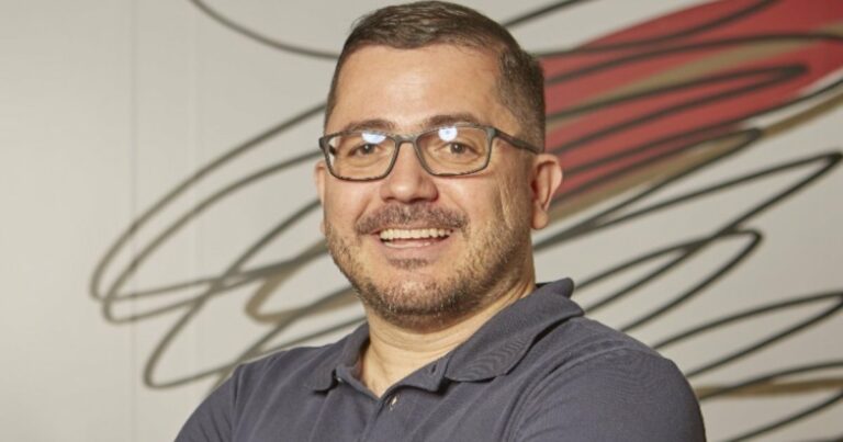 Foto de Everson Ramos, da Vórtx, sorrindo e posando com os braços cruzados. Ele usa óculos e uma camisa polo azul. O fundo apresenta um design abstrato com linhas em tons de vermelho e preto (projeto)