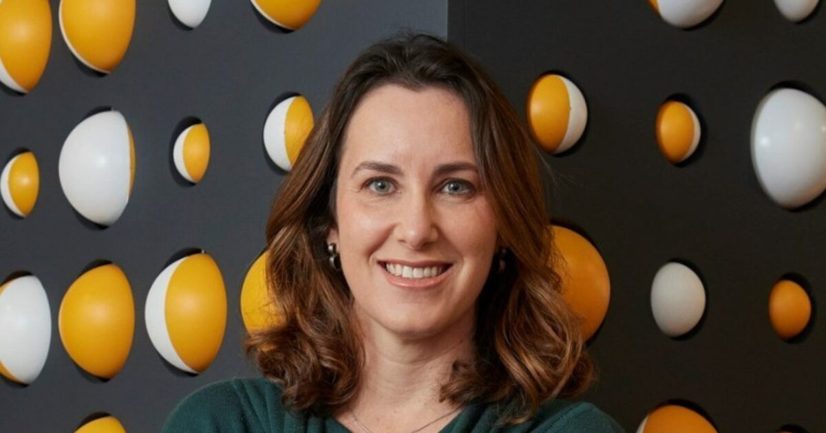 Imagem de Juliana Sztrajtman, nova country manager da Amazon Brasil, sorrindo, com cabelo castanho ondulado, vestindo roupa verde. O fundo apresenta um padrão decorativo com esferas amarelas e brancas em uma parede preta