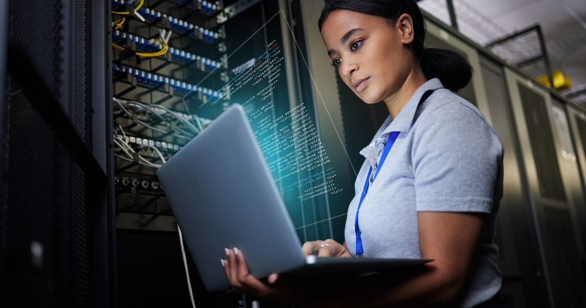 Uma mulher técnica de TI em um data center segura um laptop enquanto trabalha. Ao fundo, racks de servidores com cabos organizados estão visíveis. Um holograma com código em destaque aparece flutuando na frente do laptop, indicando trabalho relacionado a programação ou análise de dados. A profissional veste uma camisa polo cinza e um crachá, com um ambiente moderno e tecnológico ao redor (gartner, Coursera)