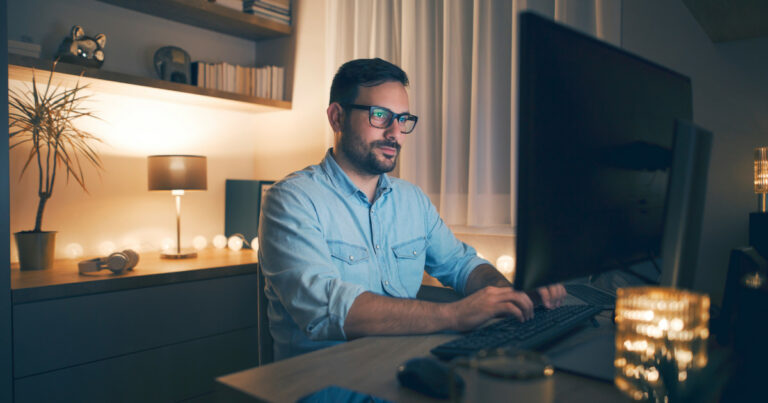 Homem usando óculos, sentado em frente a um computador, trabalhando em um ambiente aconchegante e bem iluminado. O local tem uma decoração moderna, com luminárias, plantas, estante de livros e itens decorativos ao fundo. A luz suave cria uma atmosfera confortável, ideal para trabalho remoto ou estudo (Robert Half, trabalho remoto, presencial, home office)