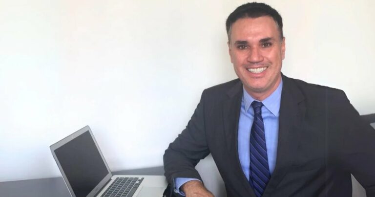 A imagem mostra Franklin Tomich, fundador da Accordia, sentado à mesa com um laptop ao lado. Ele veste um terno escuro, camisa azul e gravata listrada, transmitindo um visual profissional. Com um sorriso confiante, ele olha diretamente para a câmera. O fundo é simples, com uma parede branca, destacando o ambiente corporativo e minimalista da foto (Franklin Tomich, Accordia, risco)