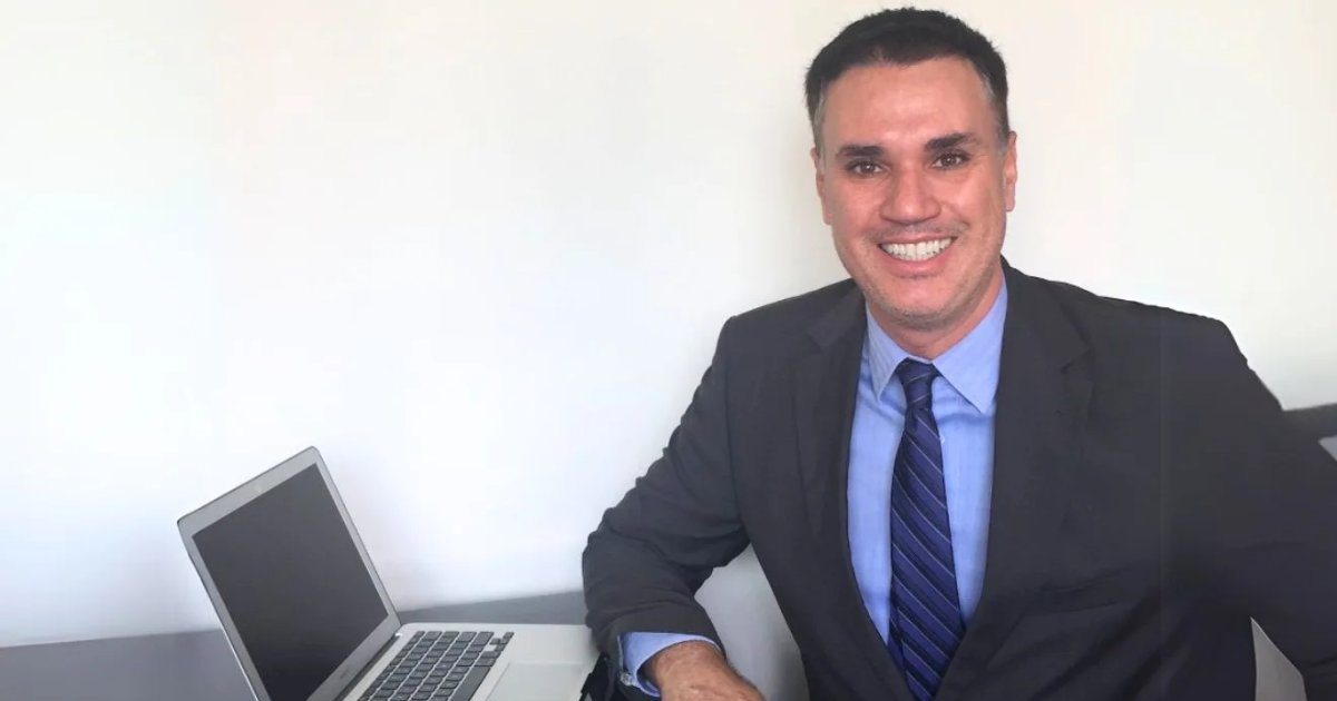 A imagem mostra Franklin Tomich, fundador da Accordia, sentado à mesa com um laptop ao lado. Ele veste um terno escuro, camisa azul e gravata listrada, transmitindo um visual profissional. Com um sorriso confiante, ele olha diretamente para a câmera. O fundo é simples, com uma parede branca, destacando o ambiente corporativo e minimalista da foto (Franklin Tomich, Accordia, risco)