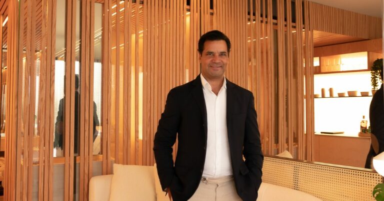 Gustavo Filgueiras, CEO do Grupo Emiliano, posa para a foto com um sorriso, vestindo um blazer preto sobre uma camisa branca e calça bege. O ambiente sofisticado e bem iluminado apresenta painéis de madeira verticalizados, decoração minimalista e tons neutros, transmitindo elegância e modernidade.