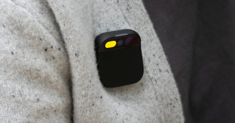 A imagem mostra um Humane Ai Pin, um dispositivo tecnológico compacto e vestível, fixado na roupa de uma pessoa. O aparelho tem um design quadrado com cantos arredondados e uma superfície preta fosca. Na parte superior, há uma seção que abriga sensores, câmeras e uma luz amarela acesa, indicando que o dispositivo está ativo. Ele está preso a um tecido de lã clara com pequenas manchas escuras, destacando seu design minimalista e discreto.