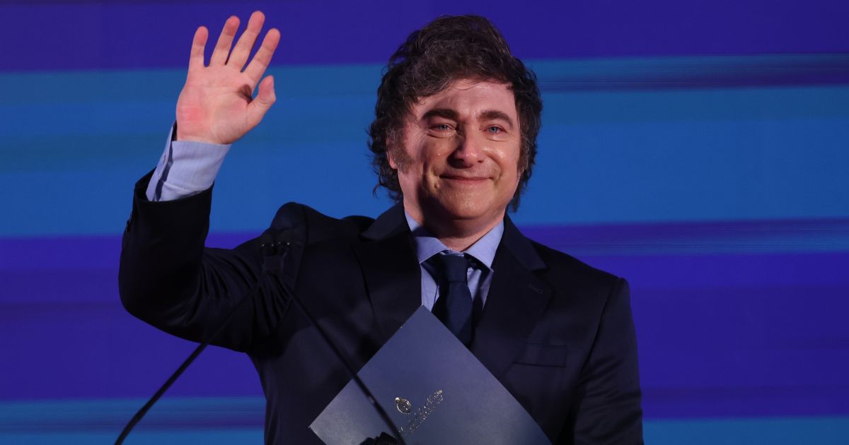 A imagem mostra um homem de terno preto, camisa azul e gravata escura, sorrindo e acenando com a mão direita levantada. Ele segura uma pasta azul com um brasão dourado. O fundo é azul com tons de luz. O homem na imagem foi identificado como Javier Milei, presidente da Argentina