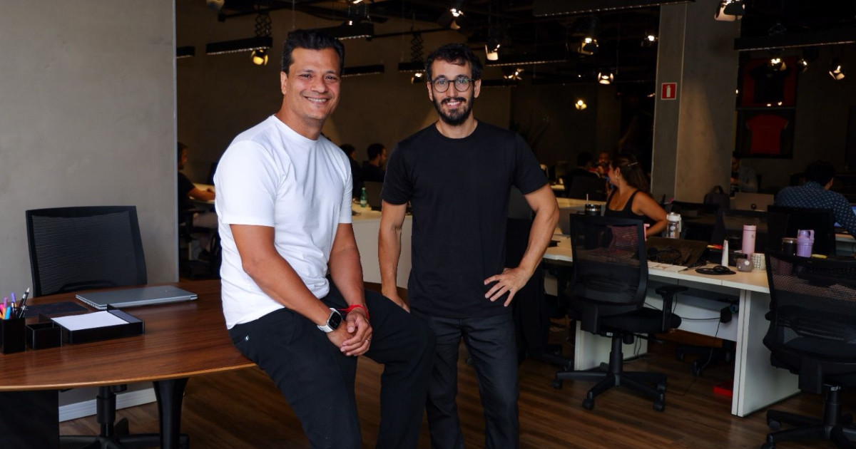 A imagem mostra Juliano Tubino, o novo CEO do grupo Alura, e Paulo Silveira, fundador da Alura e agora chief visionary officer (CVO) da organização. Eles estão em um ambiente de escritório moderno, com um fundo que inclui diversos funcionários trabalhando em suas estações. Juliano Tubino, à esquerda, veste uma camiseta branca e calça preta, sentado sobre uma mesa de madeira. Paulo Silveira, à direita, veste uma camiseta preta e calça escura, de pé ao lado dele. Ambos sorriem para a câmera, transmitindo um clima descontraído e profissional