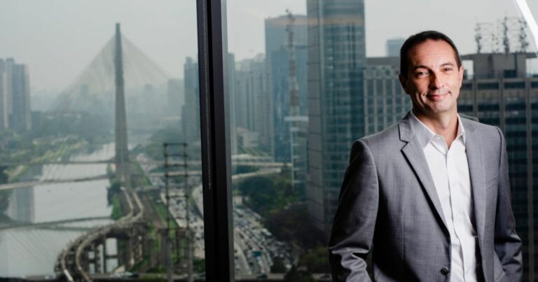 Marcelo Tangione, presidente da Mastercard Brasil, posa em um escritório com vista panorâmica para a cidade de São Paulo, destacando ao fundo a Ponte Estaiada e os arranha-céus da região.