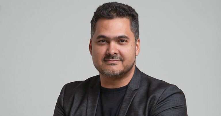 Foto de um homem de pele clara, barba curta e cabelo escuro levemente ondulado. Ele veste um blazer preto sobre uma camisa preta e tem uma expressão confiante e amigável. Seus braços estão cruzados e o fundo é um cinza neutro, conferindo um aspecto profissional à imagem (token)