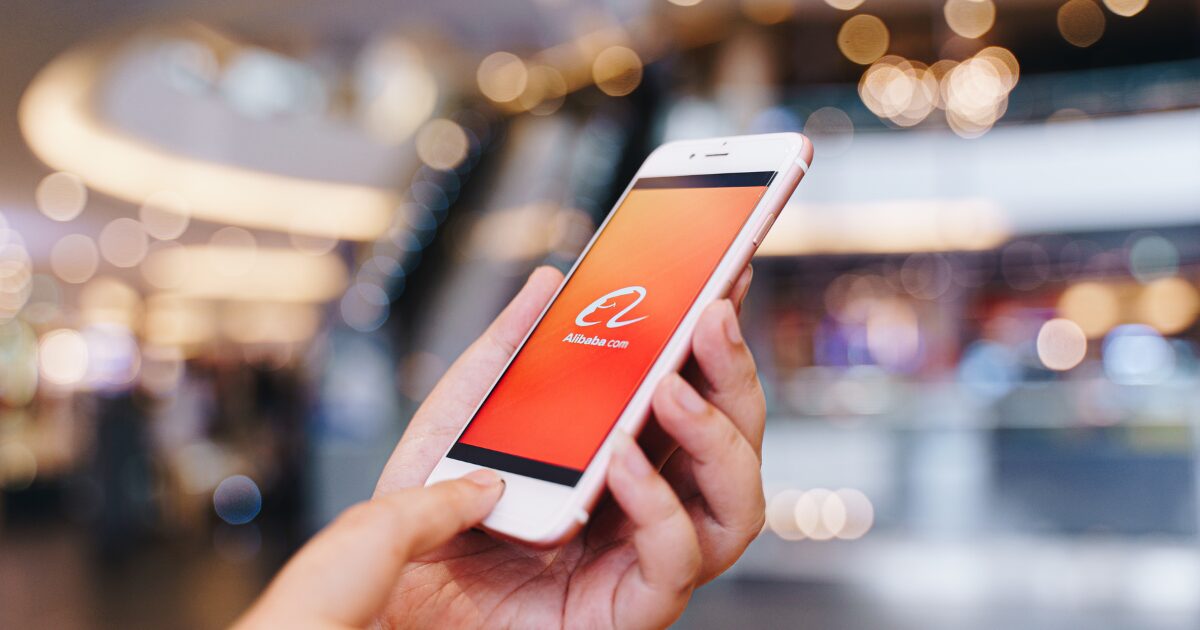 Uma pessoa segura um iPhone da Apple em um ambiente iluminado, provavelmente um shopping center. Na tela do smartphone, está aberto o aplicativo do Alibaba.com, com seu icônico logotipo branco sobre um fundo laranja. O fundo da imagem está desfocado, criando um efeito de luzes circulares, enquanto o foco principal está no dispositivo e na interação do usuário com a tela