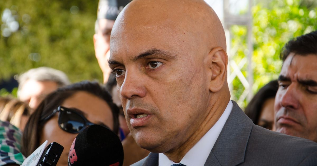 A imagem mostra um homem calvo, de pele clara, vestindo um terno cinza, camisa branca e gravata azul. Ele tem uma expressão séria e parece estar falando com jornalistas, pois há diversos microfones à sua frente, incluindo um da "Rádio Bandeirantes". Atrás dele, há várias pessoas, algumas usando óculos escuros e outras com expressões atentas ou sérias. O fundo é externo, com árvores verdes e uma estrutura metálica visível. A cena transmite a sensação de uma entrevista coletiva ou uma declaração pública. Rumble