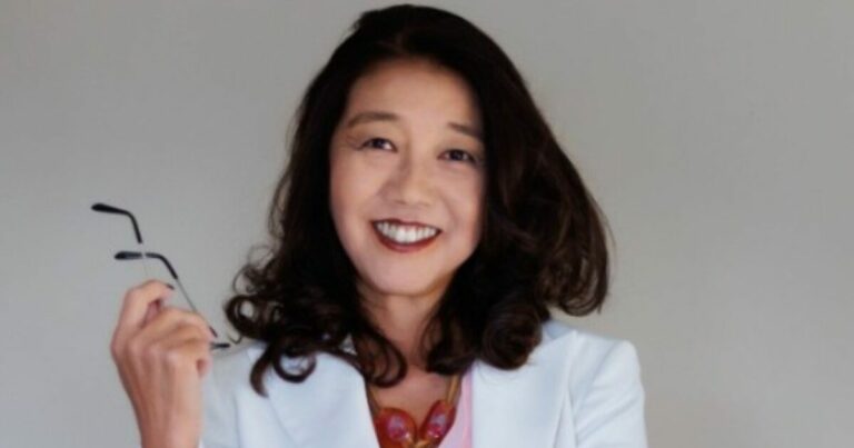 Nancy Abe sorridente de cabelos castanhos longos e ondulados, vestindo um blazer branco sobre uma blusa rosa e um colar de pedras vermelhas. Ela segura um par de óculos na mão direita e olha diretamente para a câmera com expressão confiante. O fundo é neutro e cinza claro.