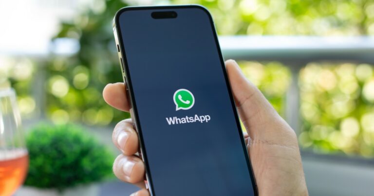 A imagem mostra uma mão segurando um smartphone com a tela ligada, exibindo a tela de carregamento do aplicativo WhatsApp. O logotipo verde e branco do WhatsApp está centralizado na tela preta, com o nome do aplicativo logo abaixo. O fundo da imagem está levemente desfocado, mostrando um ambiente ao ar livre com plantas verdes e um copo com bebida alaranjada ao lado