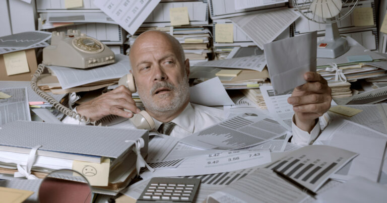 A imagem mostra um homem de meia-idade, de aparência exausta, vestindo uma camisa branca e gravata, cercado por uma enorme quantidade de papéis, documentos, gráficos e arquivos. Ele está ao telefone com uma expressão de cansaço e confusão, enquanto segura um envelope ou ficha com a outra mão. O ambiente parece ser um escritório caótico, com uma mesa e prateleiras desorganizadas, lotadas de papéis, pastas e notas adesivas. Ao fundo, um telefone antigo, um ventilador e mais pilhas de documentos reforçam a sensação de desordem extrema (caos)