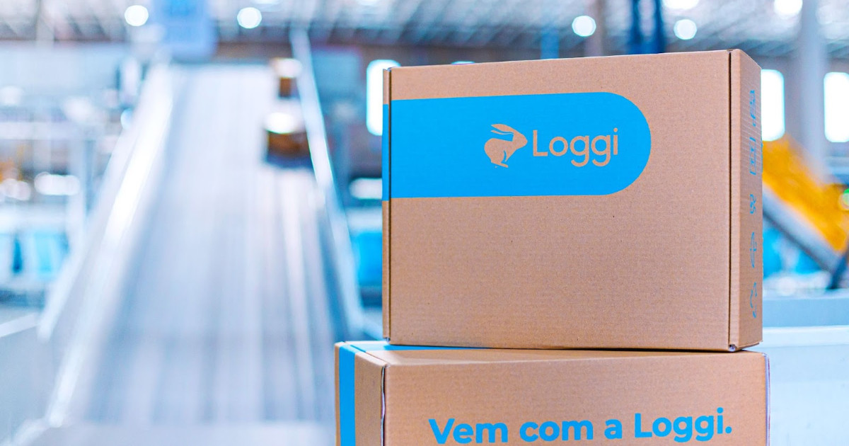 Caixas de encomenda da Loggi em destaque, com a logo da empresa e a frase 'Vem com a Loggi' impressas. O fundo desfocado mostra um centro de distribuição automatizado, com esteiras e iluminação moderna, representando a logística eficiente e tecnologia aplicada ao transporte de mercadorias (plataformas)