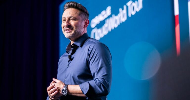 Alexandre Maioral, presidente da Oracle do Brasil, está em pé no palco durante um evento. Ele sorri enquanto segura um controle nas mãos. Ao fundo, há uma grande tela azul com seu nome e cargo exibidos: "Alexandre Maioral, Presidente, Oracle do Brasil".