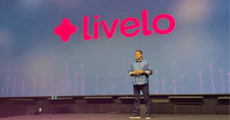 CEO da Livelo em pé no palco durante apresentação corporativa. Ele veste camisa jeans e calça escura, com o logotipo da Livelo projetado em grande escala no telão atrás dele. A iluminação do palco é predominantemente rosa e roxa, com detalhes em verde neon nas bordas do palco.