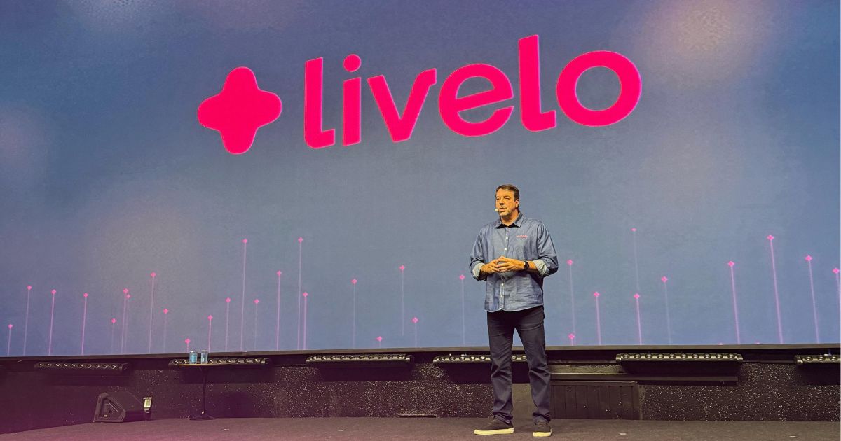CEO da Livelo em pé no palco durante apresentação corporativa. Ele veste camisa jeans e calça escura, com o logotipo da Livelo projetado em grande escala no telão atrás dele. A iluminação do palco é predominantemente rosa e roxa, com detalhes em verde neon nas bordas do palco.
