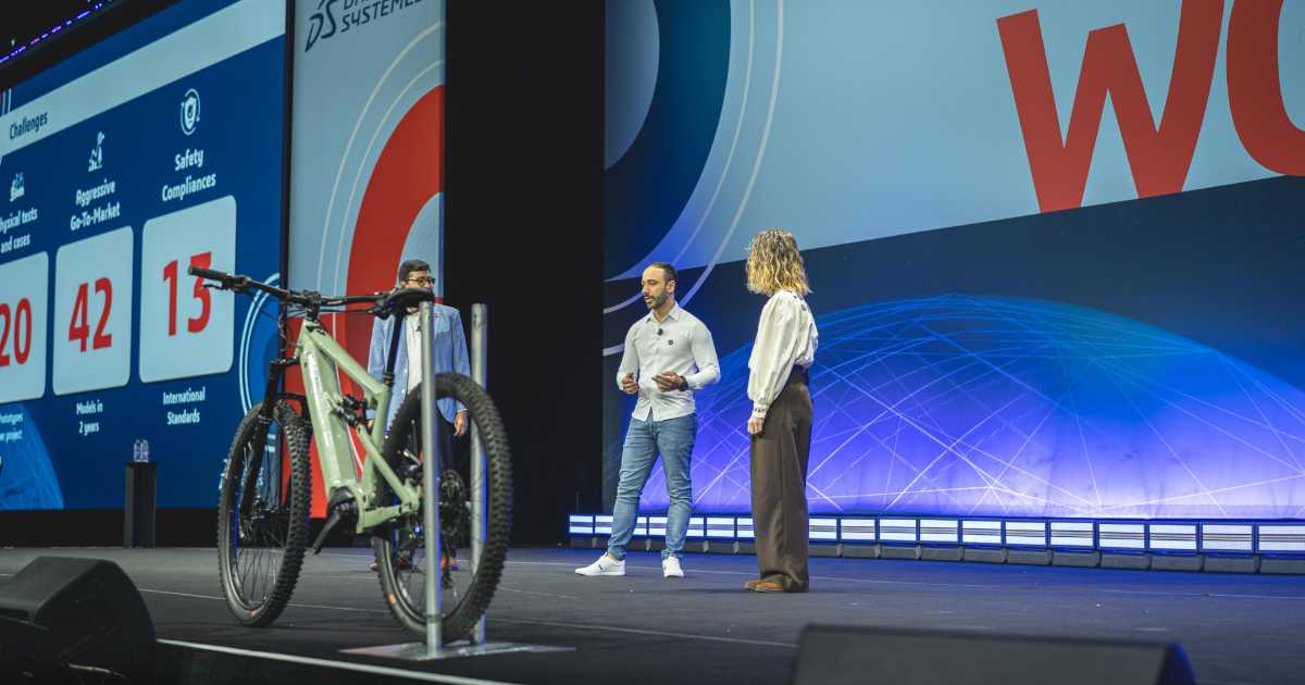 Leandro Timotio da Silva, engenheiro de produtos da Caloi, apresenta-se no palco do 3DExperience World 2025, evento da Dassault Systèmes. Ele está acompanhado de duas outras pessoas, uma mulher e um homem, enquanto fala ao público. No palco, há uma bicicleta exposta em destaque, e ao fundo, um grande telão exibe informações sobre desafios da empresa, incluindo números sobre testes físicos, modelos desenvolvidos e padrões de segurança.