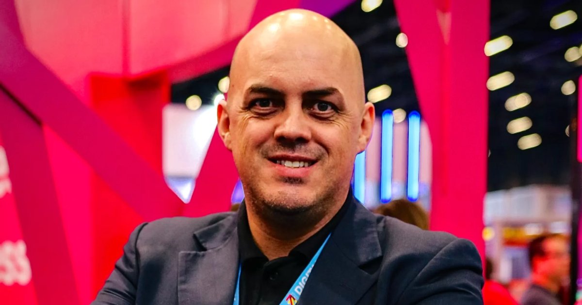 Homem de pele clara, calvo, com barba rala, sorrindo, veste paletó escuro e camiseta preta. Ao fundo, ambiente interno com iluminação artificial e estruturas nas cores rosa e azul, possivelmente em um evento corporativo. A imagem mostra Daniel Sartori, Head de Conectividade Móvel no Brasil da Deutsche Telekom Global Business (Brasil)