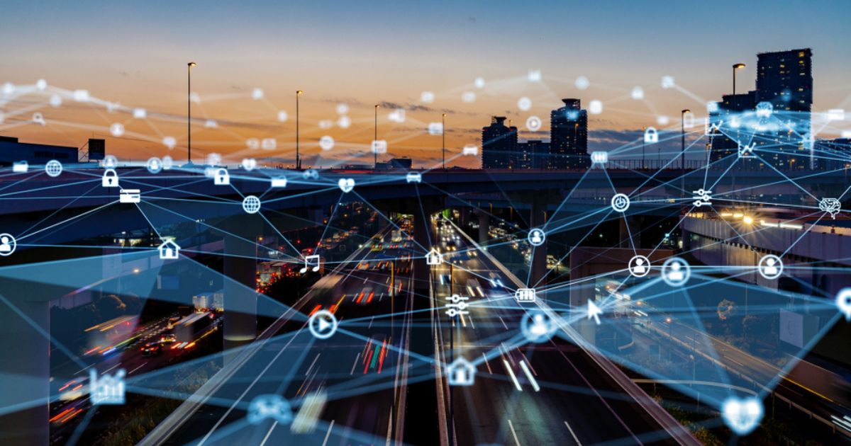 Imagem de uma cidade inteligente ao entardecer, com rodovias movimentadas e prédios modernos ao fundo. Sobre a cena urbana, há uma sobreposição de elementos gráficos representando conexões digitais e a infraestrutura de edge computing. Ícones de dispositivos conectados, como nuvem, cadeados, casas e globos, estão interligados por linhas, simbolizando a descentralização do processamento de dados na borda da rede. A composição visual reforça o conceito de conectividade avançada e Internet das Coisas (IoT) em um ambiente urbano digitalizado.