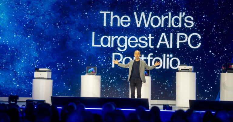 A imagem mostra Alex Cho, presidente de Personal Systems da HP, no palco durante um evento da HP Amplify. Ele está apresentando novos produtos da linha de PCs com inteligência artificial, com um grande telão ao fundo exibindo a frase "The World's Largest AI PC Portfolio" ("O maior portfólio de PCs com IA do mundo"). No palco, há pedestais com notebooks, impressoras e outros dispositivos da HP. A plateia aparece na parte inferior da imagem, assistindo à apresentação