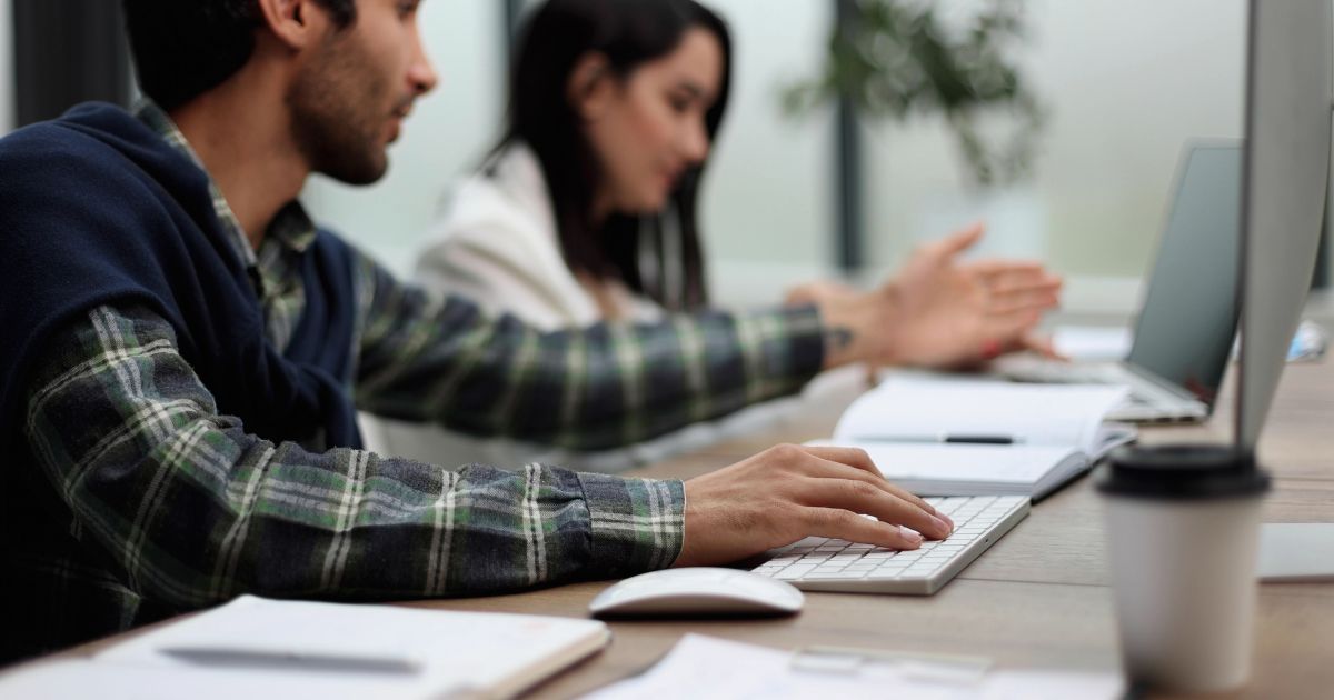 A imagem mostra duas pessoas trabalhando em um ambiente de escritório moderno. No primeiro plano, um homem de camisa xadrez verde digita em um teclado enquanto olha para o ecrã do computador. Ao fundo, uma mulher de cabelo escuro, vestindo um blazer claro, parece estar envolvida em uma conversa ou explicação, gesticulando com a mão. A mesa contém vários objetos, incluindo um copo de café descartável, cadernos e laptops. O ambiente tem uma estética contemporânea, com iluminação natural vinda de grandes janelas.