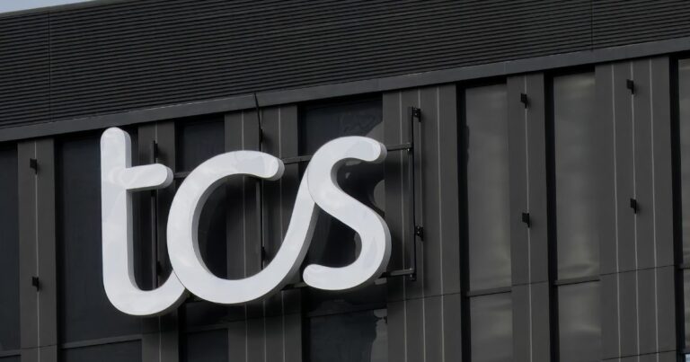 Um letreiro iluminado com as letras "tcs" em branco está fixado na fachada de um prédio moderno de cor escura. A estrutura do edifício apresenta janelas verticais e detalhes arquitetônicos minimalistas. O céu ao fundo está parcialmente nublado, refletindo sutilmente na superfície do letreiro (mulher)