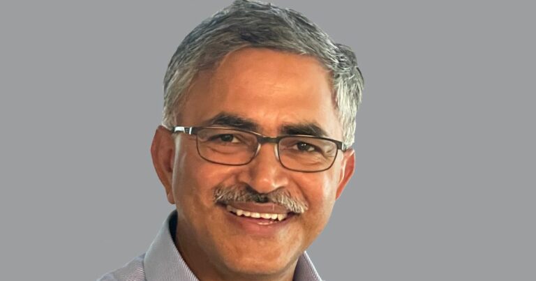 Um homem de meia-idade, com cabelos grisalhos curtos e bigode, está sorrindo. Ele usa óculos de armação fina e uma camisa social de cor clara. O fundo da imagem é neutro e sólido, destacando o rosto do homem. O homem é Hasmukh Ranjan, CIO global da AMD.