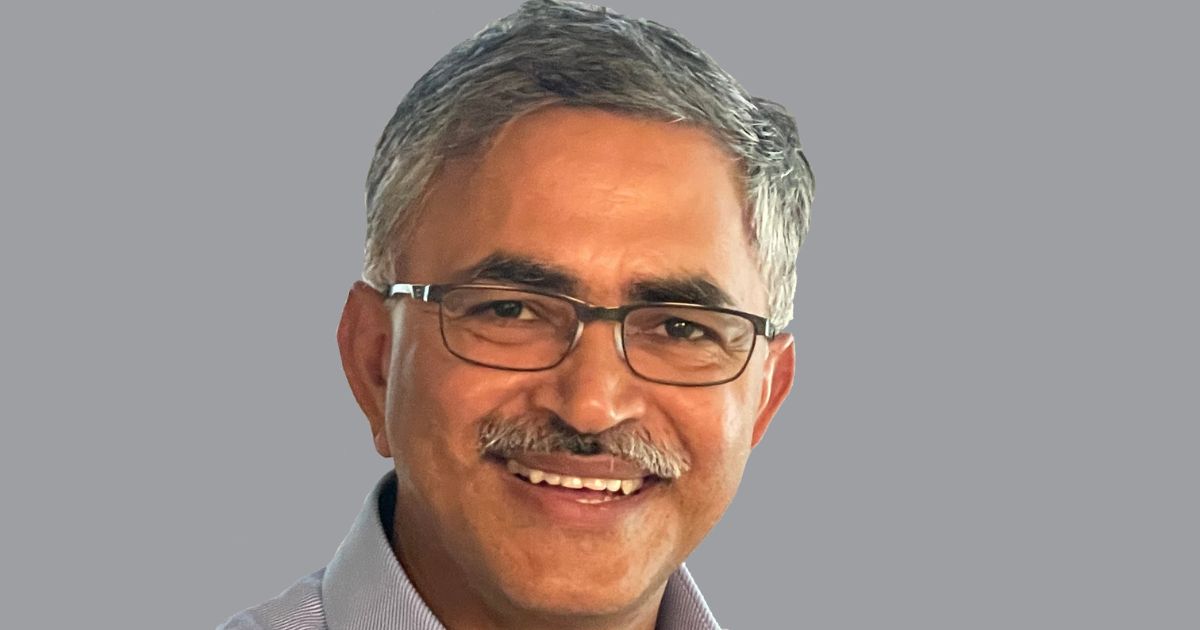 Um homem de meia-idade, com cabelos grisalhos curtos e bigode, está sorrindo. Ele usa óculos de armação fina e uma camisa social de cor clara. O fundo da imagem é neutro e sólido, destacando o rosto do homem. O homem é Hasmukh Ranjan, CIO global da AMD.