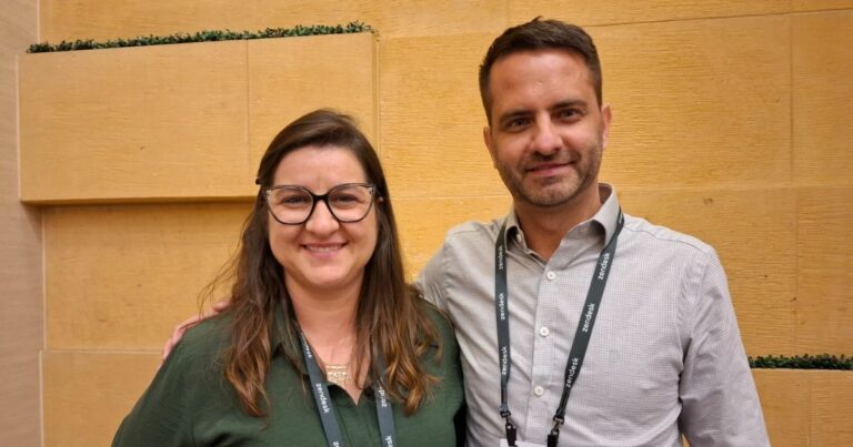Um homem e uma mulher estão juntos, sorrindo para a câmera. A mulher tem cabelos castanhos longos, usa óculos de armação preta e uma blusa verde. O homem tem cabelos curtos, barba curta e usa uma camisa social cinza. Ambos têm crachás pendurados no pescoço e estão em frente a uma parede de tom bege com pequenos detalhes verdes no topo.