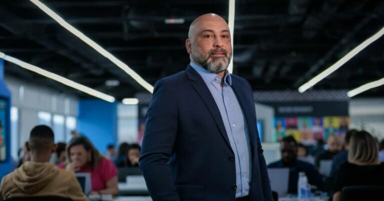 Um retrato em plano médio de um homem careca de barba grisalha, vestido com um blazer azul-marinho e uma camisa azul clara. Ele está posando em um escritório moderno com outros indivíduos trabalhando em segundo plano. A iluminação do escritório é brilhante, com luminárias lineares no t
