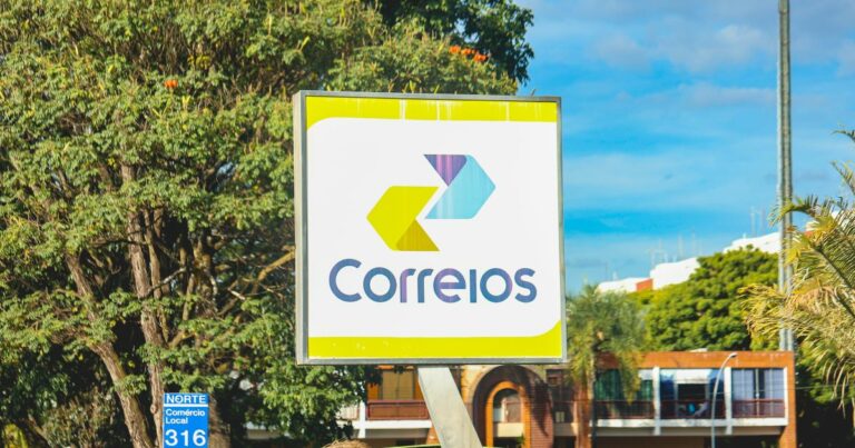 A imagem mostra uma placa dos Correios do Brasil em um ambiente urbano. O letreiro exibe o logotipo oficial da empresa, composto por duas setas estilizadas nas cores amarela e azul, apontando para a direita, junto com a palavra "Correios" escrita em letras azuis. A placa tem uma moldura metálica e fundo branco com detalhes em amarelo. No canto inferior esquerdo da imagem, há uma pequena placa azul com a inscrição "NORTE Comércio Local 316". Ao fundo, é possível ver árvores, um céu azul com algumas nuvens e edifícios com arquitetura típica de áreas comerciais e residenciais.