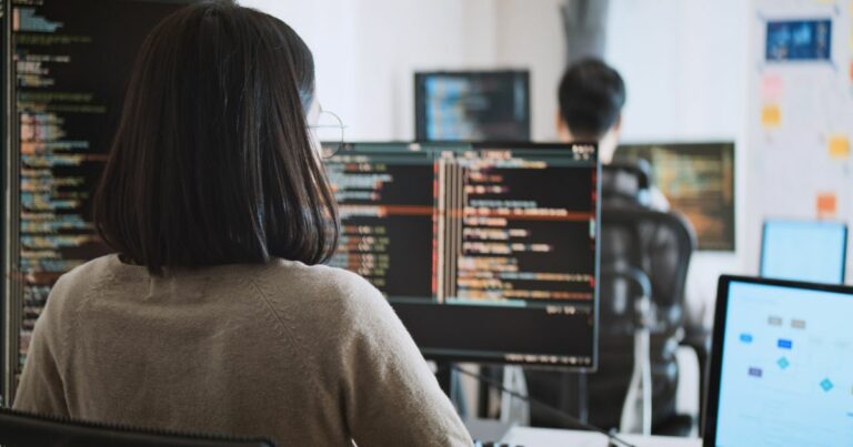 Uma mulher de cabelo escuro e óculos está sentada em frente a vários monitores de computador, trabalhando com código de programação. A tela principal exibe linhas de código coloridas em um ambiente de desenvolvimento. No fundo, outro programador, um homem de cabelo curto, também trabalha em seu computador, e há um quadro branco com anotações e diagramas. O ambiente parece ser um escritório moderno de tecnologia ou uma startup. (contrata)