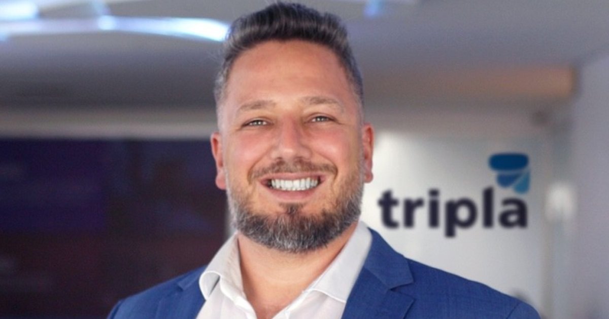 Homem de pele clara, barba e cabelo castanho-escuros com topete, sorrindo, veste paletó azul e camisa social branca. Ao fundo, ambiente corporativo com o logotipo da empresa "Tripla" na parede. A imagem mostra Rodrigo Colen, sócio-fundador e Diretor Comercial da Tripla (202)