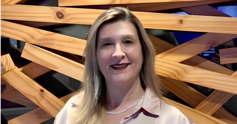 Tamara Gussiardi, head de TI da Nestlé Brasil, sorri em frente a um painel decorativo de ripas de madeira entrelaçadas. Ela veste uma camisa de seda clara e colar discreto, com cabelos loiros soltos.