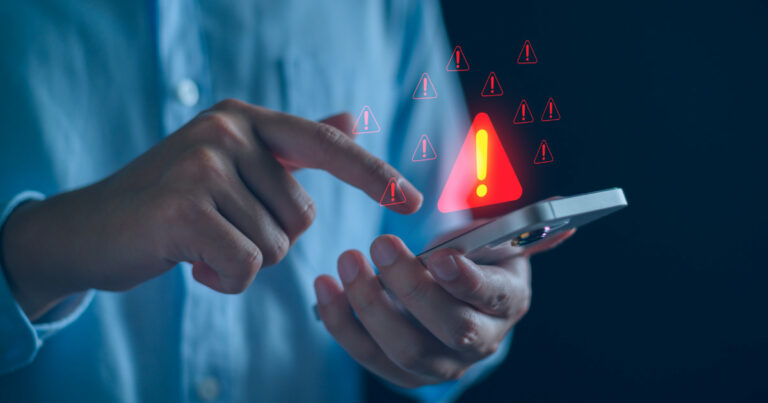 A imagem mostra uma pessoa segurando um smartphone enquanto toca na tela com o outro dedo. Diversos ícones vermelhos de alerta com pontos de exclamação aparecem sobrepostos à cena, simbolizando um aviso ou risco relacionado ao uso do dispositivo. O fundo escuro e desfocado destaca a interação e transmite uma sensação de preocupação ou atenção necessária, sugerindo um possível problema de segurança digital, golpe ou notificação de erro (avisamos)