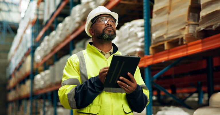 Homem vestindo capacete branco e jaqueta refletiva amarela segura um tablet enquanto realiza inspeção ou controle de estoque dentro de um armazém logístico. Ao fundo, prateleiras altas armazenam caixas e produtos organizados, representando o conceito de cadeia de suprimentos (indústria)