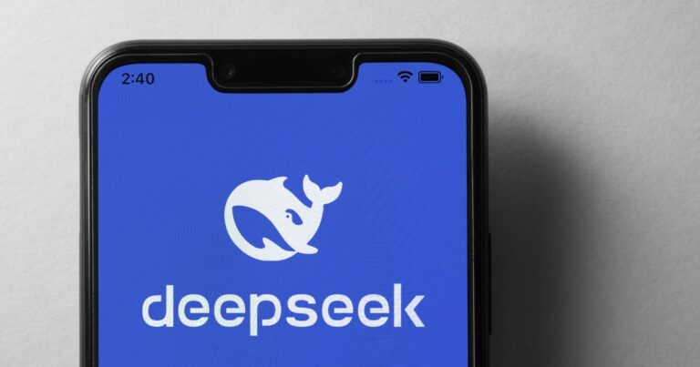 A imagem mostra um smartphone com a tela exibindo o logotipo da "deepseek" sobre um fundo azul. O logotipo consiste em uma baleia estilizada em branco, acompanhada do nome da empresa em letras minúsculas. O aparelho tem um design moderno, com bordas finas e um entalhe na parte superior da tela. A imagem transmite uma estética tecnológica e minimalista, possivelmente relacionada a inteligência artificial, pesquisa ou desenvolvimento de software, microsoft
