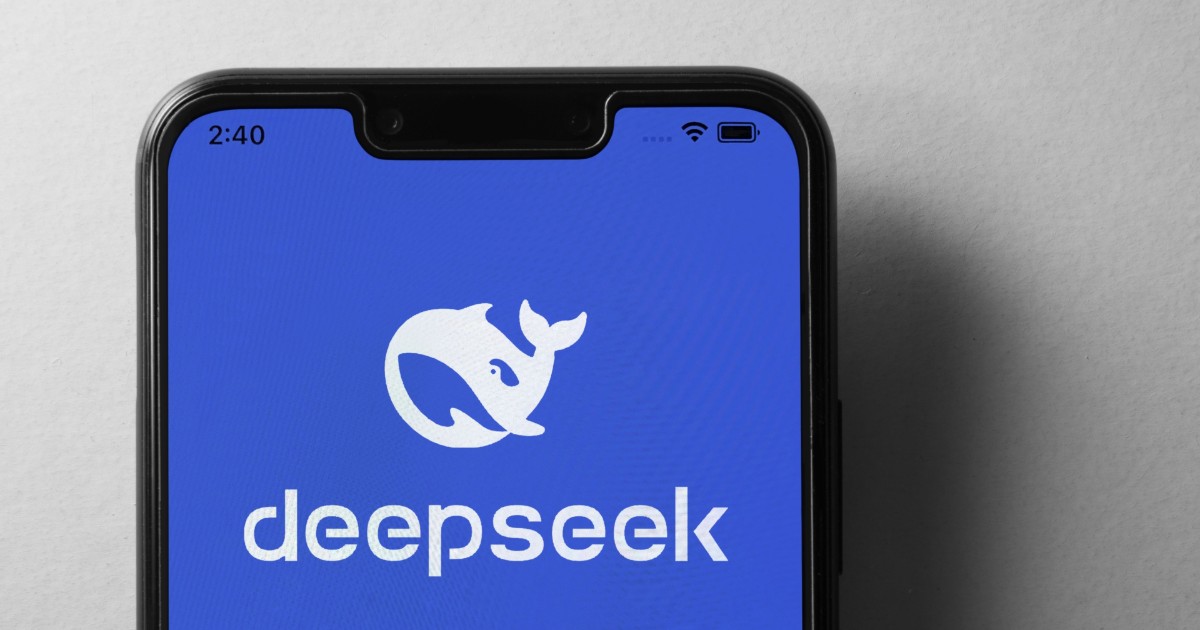 A imagem mostra um smartphone com a tela exibindo o logotipo da "deepseek" sobre um fundo azul. O logotipo consiste em uma baleia estilizada em branco, acompanhada do nome da empresa em letras minúsculas. O aparelho tem um design moderno, com bordas finas e um entalhe na parte superior da tela. A imagem transmite uma estética tecnológica e minimalista, possivelmente relacionada a inteligência artificial, pesquisa ou desenvolvimento de software, microsoft