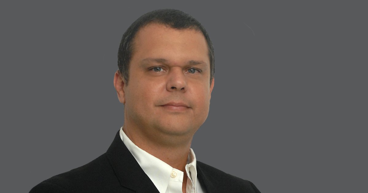 Homem de pele clara, cabelo curto e escuro, vestindo paletó preto e camisa social branca, posando de frente com expressão séria. Fundo cinza liso. A imagem mostra Marcelo Rodrigues, country manager da Commvault
