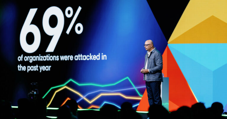 Um homem careca, de óculos e vestindo casaco cinzento e calças escuras, está em pé sobre um palco a fazer uma apresentação. Atrás dele, há um ecrã gigante com gráficos coloridos e texto em inglês que diz: “69% of organizations were attacked in the past year” (“69% das organizações foram atacadas no último ano”). A plateia está em silhueta no primeiro plano, observando atentamente. O cenário tem elementos gráficos coloridos em azul, vermelho, laranja e amarelo, criando um fundo moderno e tecnológico (Anand Eswaran, CEO da Veeam)