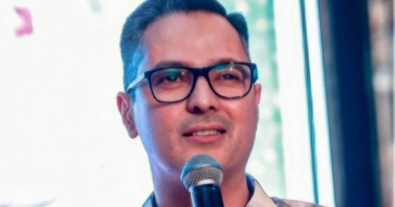 Eronides Junior, novo Chief Revenue Officer (CRO) da SoftwareOne, aparece segurando um microfone e sorrindo, usando óculos de grau e camisa clara, com fundo desfocado e iluminação suave.