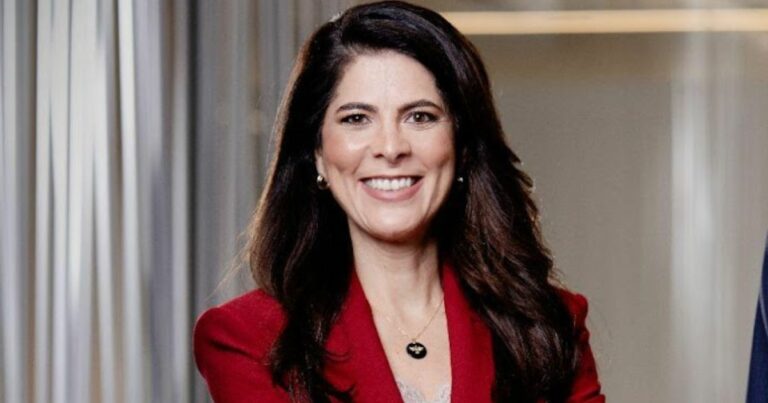 Imagem de Roberta Godoi, CEO da Claro Empresas, sorrindo. Ela tem cabelos castanhos escuros, longos e ondulados, e veste um blazer vermelho. Usa brincos discretos e um colar com pingente redondo preto. O fundo é neutro, com cortinas claras e uma parede bege.