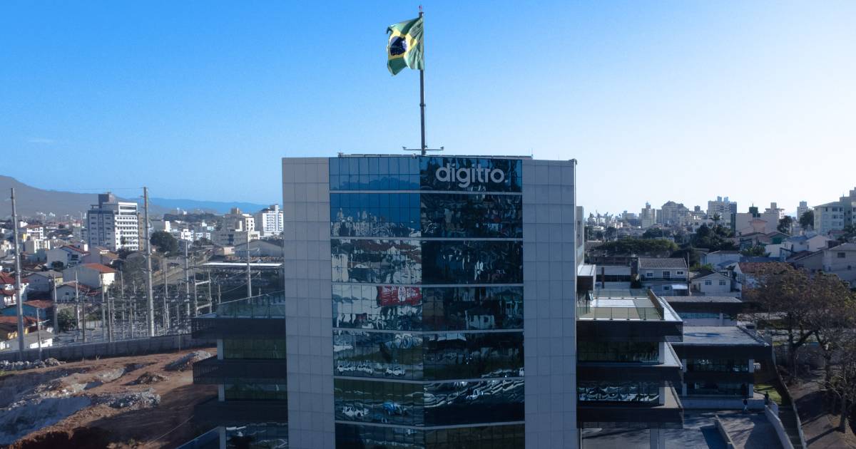 Vista superior da fachada do prédio da empresa Dígitro Tecnologia, com destaque para a logo da empresa no topo e a bandeira do Brasil hasteada em um mastro central. A fachada é composta por painéis de vidro azul escuro e estrutura lateral em tom claro, contrastando com o céu azul limpo ao fundo.