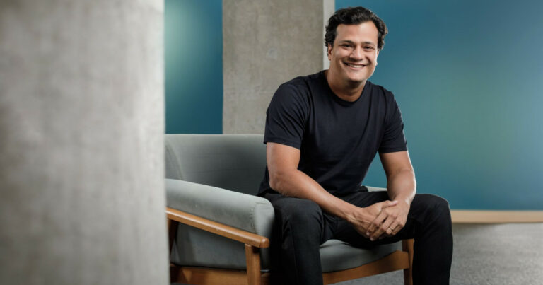 Homem sorridente, de cabelos curtos e escuros, vestindo camiseta preta e calça preta, sentado em uma poltrona cinza clara com estrutura de madeira. Ao fundo, há paredes de concreto e uma parede azul. A imagem apresenta Juliano Tubino, novo CEO do Grupo Alura.
