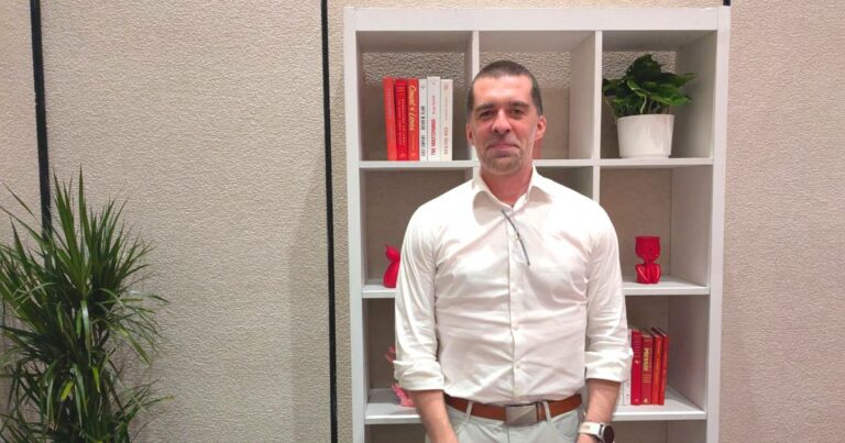 Homem em pé, usando camisa social branca e calça clara, com óculos pendurado na gola da camisa. Ao fundo, há uma estante branca com livros, objetos decorativos vermelhos e um vaso de planta. À esquerda da imagem, há uma planta decorativa. Trata-se de Mat Velloso, VP de Produto - Plataforma de Desenvolvimento em IA do Google DeepMind