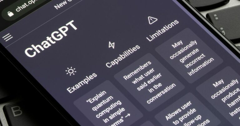 Ecrã de um smartphone com a interface do ChatGPT aberta, mostrando exemplos, capacidades e limitações da ferramenta, com um teclado de computador visível ao fundo, openai
