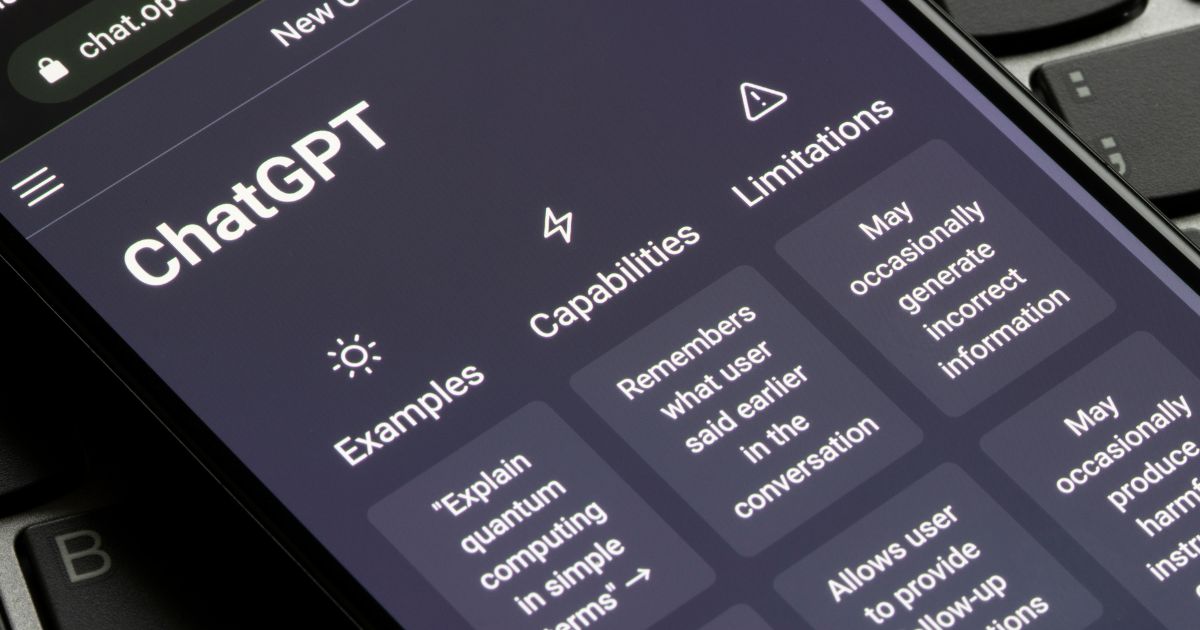Ecrã de um smartphone com a interface do ChatGPT aberta, mostrando exemplos, capacidades e limitações da ferramenta, com um teclado de computador visível ao fundo, openai