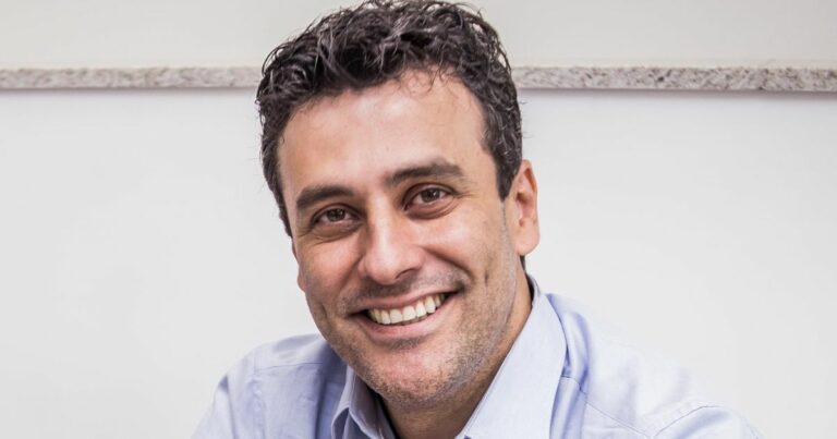 Homem sorridente com cabelo escuro e encaracolado, vestindo camisa azul clara, posando diante de uma parede branca com uma superfície de granito ao fundo. A expressão facial transmite simpatia e cordialidade.