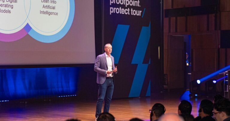 Homem a fazer uma apresentação num palco durante o evento "Proofpoint Protect Tour". Ele está vestido com blazer, camisa branca e jeans, segurando um comando. À sua esquerda há um ecrã com gráficos e textos relacionados com inteligência artificial. O público está sentado a assistir, e o ambiente é profissional e moderno, com iluminação azul. Nate Chessin, Vice-presidente sênior de engenharia de sistemas mundiais da Proofpoint